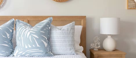A neatly made bed with a white comforter and blue patterned pillows, a wooden nightstand with a white lamp, and woven wall decorations.