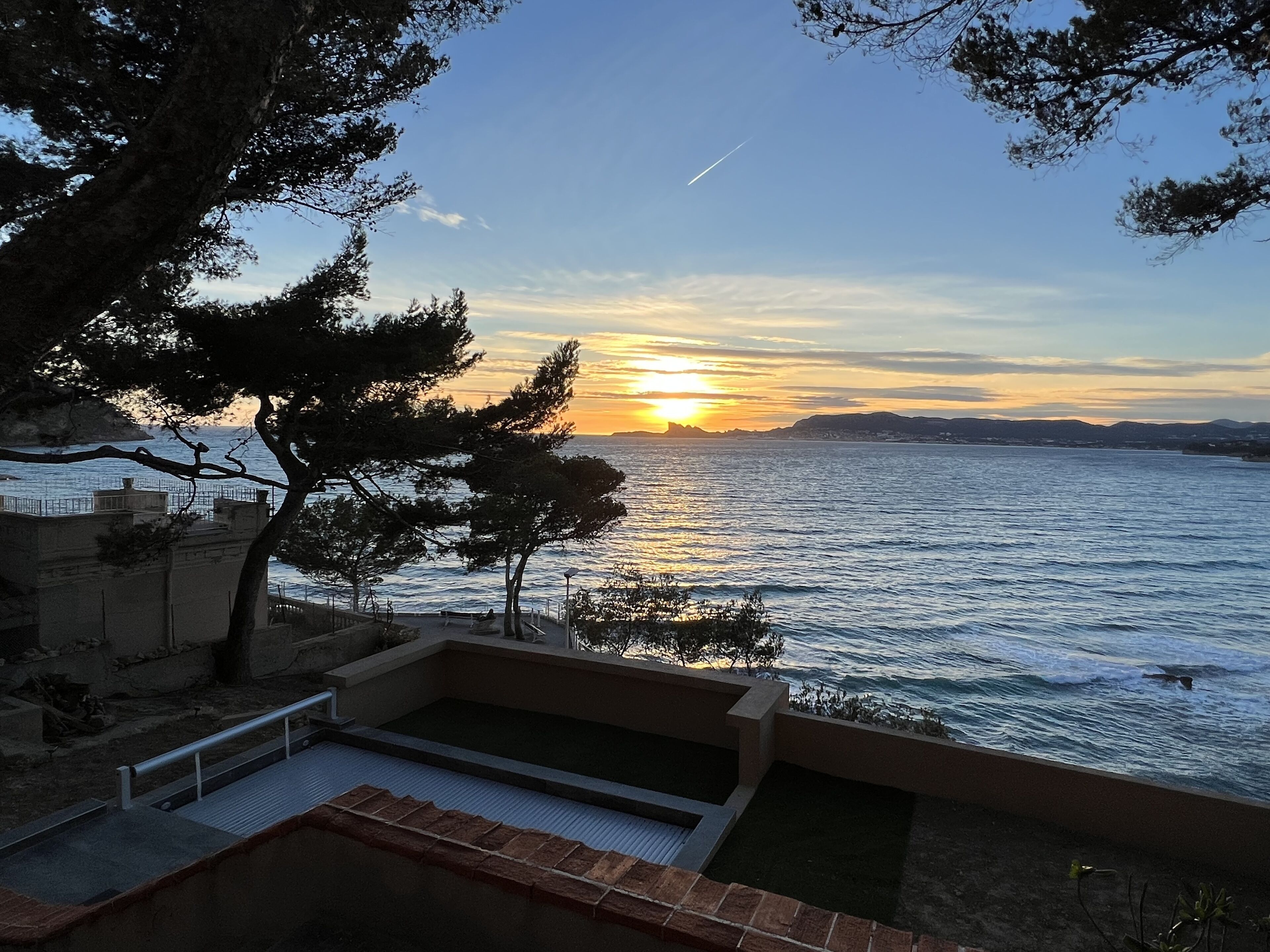 A sunset view from a balcony overlooking a body of water, with trees and a pool in the foreground.