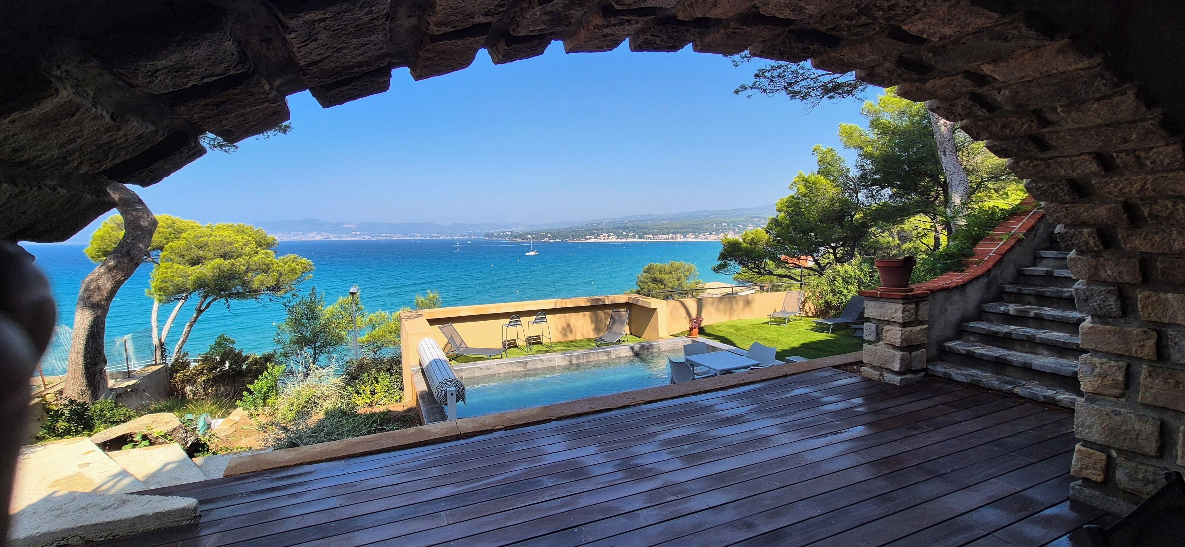 A stone archway leading to a wooden deck overlooking a pool and a coastal view.