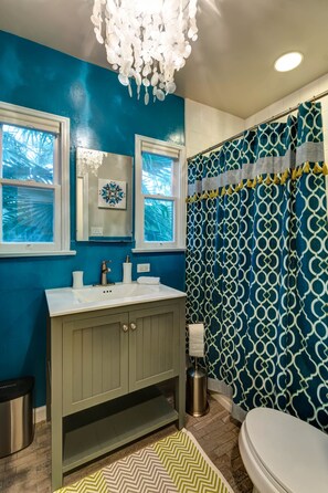 Ein Badezimmer mit blauer Wand, Waschbecken in Weiß, einem Spiegel und einer Duschvorhang mit geometrischem Muster.