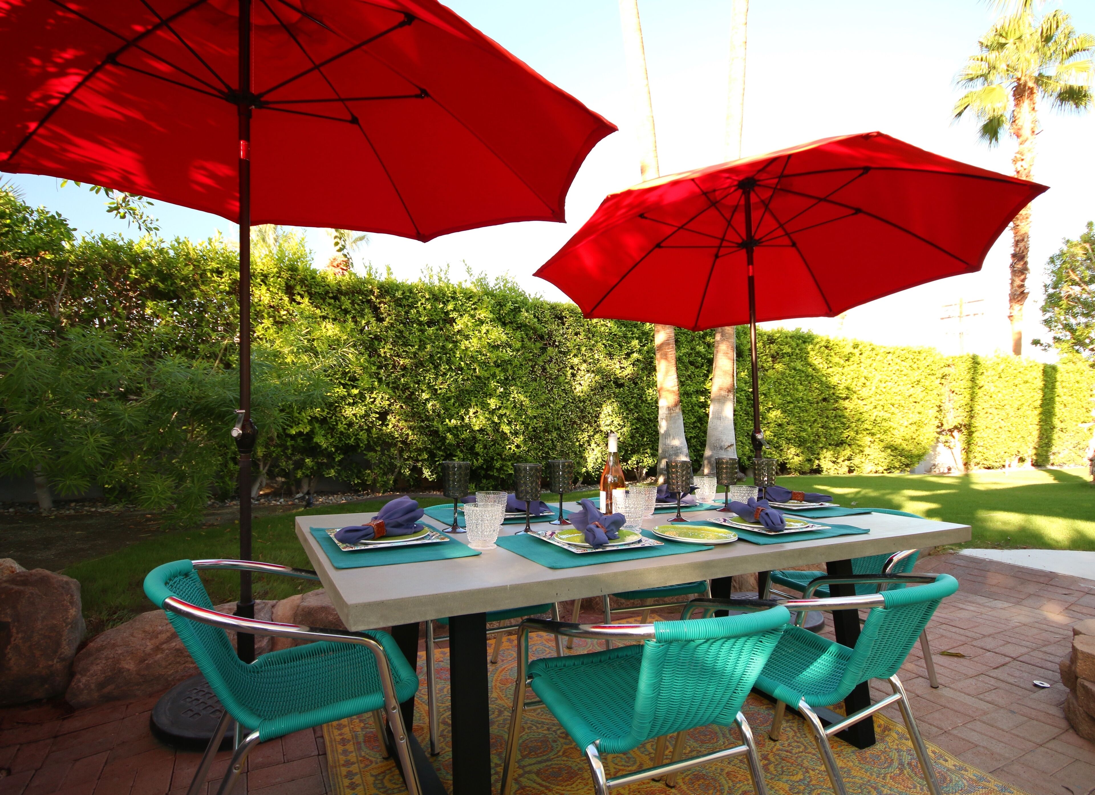 A table set outdoors with red umbrellas, green chairs, and a neatly arranged tablecloth.