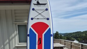 A white paddleboard with red and blue stripes, a logo, and a website name, standing on a paved deck with a wooden fence and a wooden deck in the background. - Great cabin in a central location (Agder)