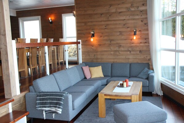 A modern living room with a grey sectional sofa, a wooden coffee table, and a view of the outdoors through large windows.