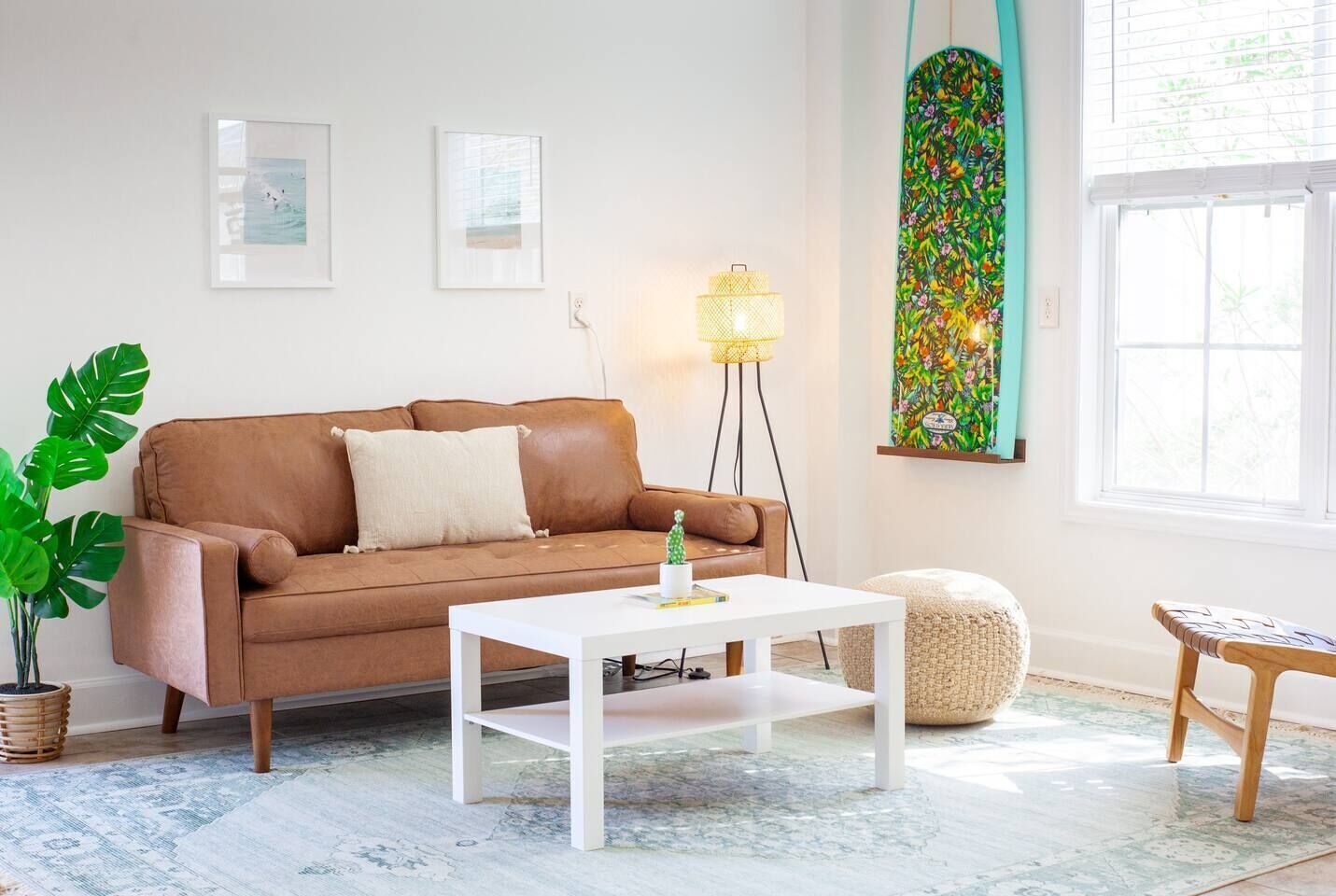 Ein Wohnzimmer mit einem braunen Ledersofa, einem weißen Couchtisch, einer Stehlampe, einem farbenfrohen Kunstwerk und einem Fenster mit Jalousien.