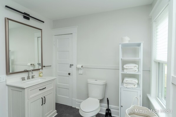 A modern bathroom with a white vanity, a large mirror, a toilet, and a window with blinds.