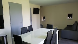 A modern living room with a white dining table, black chairs, a grey sofa, and a flat-screen TV mounted on the wall. - Petite Maison Moderne à la Campagne (Saint-Paulien)