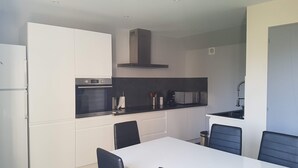 A modern kitchen with white cabinetry, a built-in oven, and a black countertop. There is a dining table with chairs and a small trash can. - Petite Maison Moderne à la Campagne (Saint-Paulien)