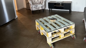 A living room with a modern kitchen, a flat-screen TV mounted on the wall, a patterned armchair, a small black console table, and a wooden pallet coffee table with wheels. - Home Away From Home (Reno)