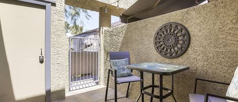 An outdoor patio with a table and two chairs, a decorative wall piece, and a door with a keypad lock.