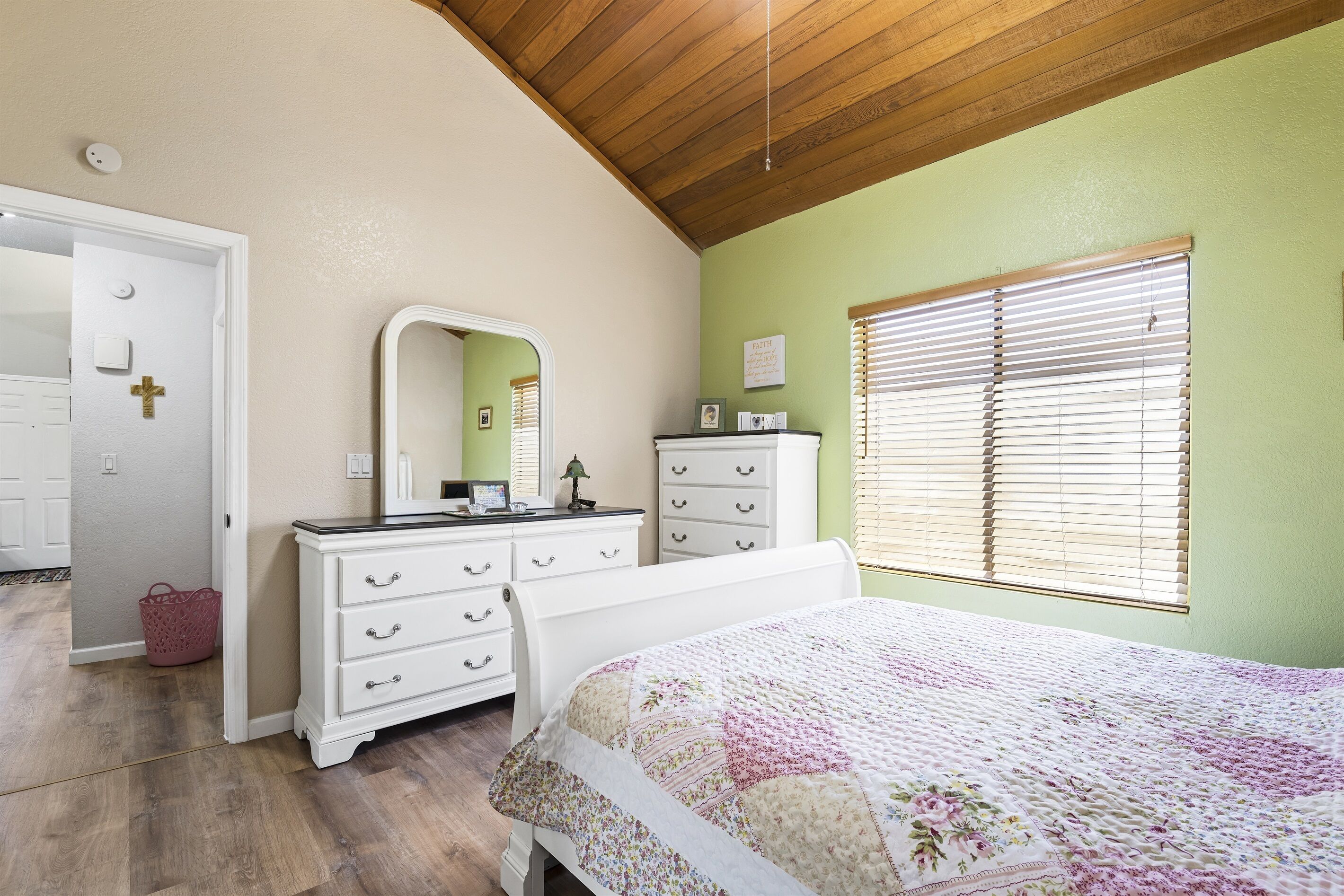 Une chambre avec un plafond en bois, un lit recouvert d’une courtepointe à motifs floraux, une commode avec des tiroirs, un miroir et une fenêtre avec des stores.
