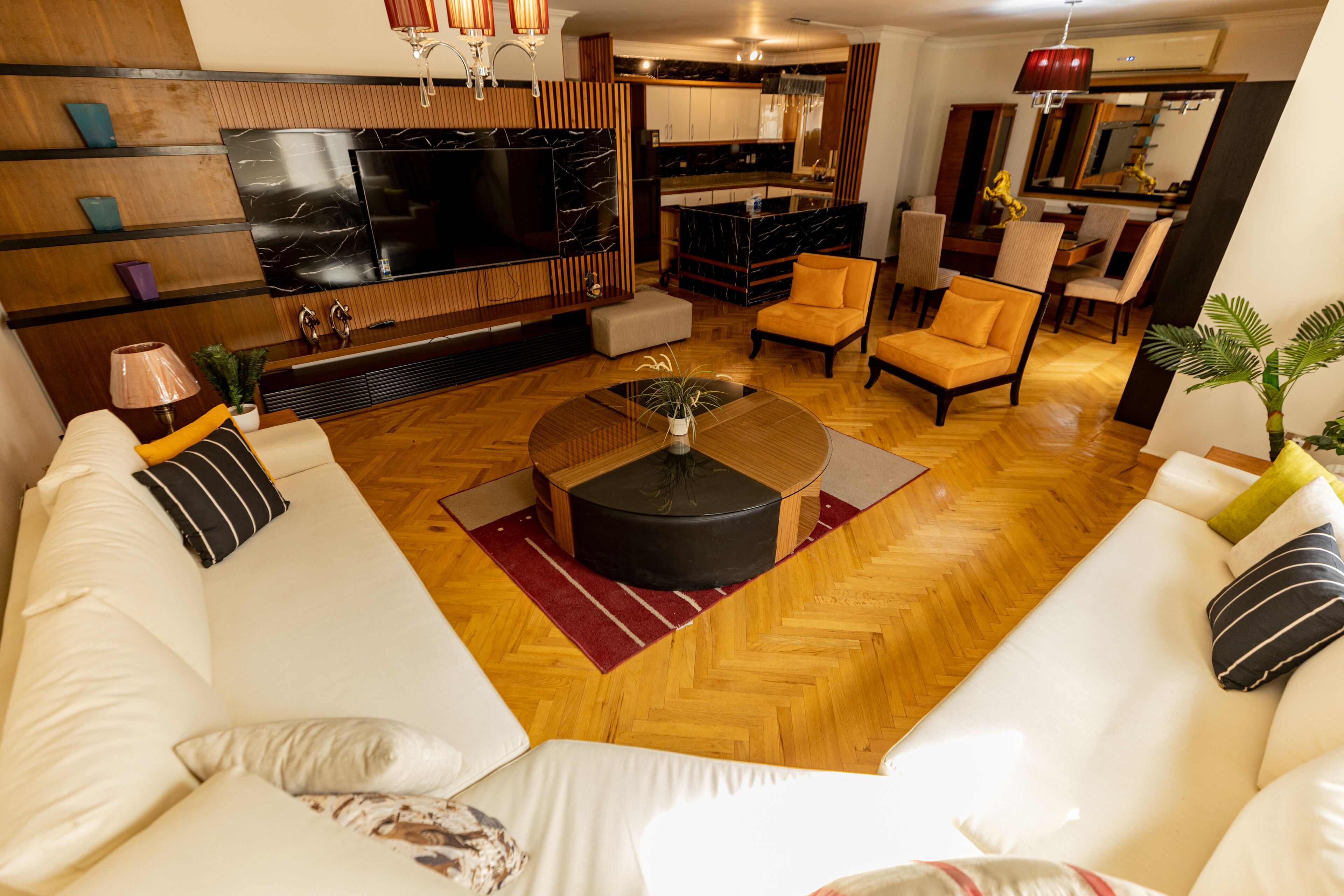 Un salon moderne doté d’un grand téléviseur à écran plat, d’un parquet en bois disposé en chevrons, d’un canapé blanc et d’un coin repas avec une cuisine en arrière-plan.
