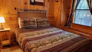 A wooden cabin room with a bed, a nightstand, a Texas flag, and a window with curtains.