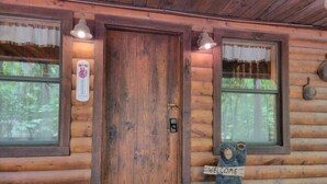 A wooden cabin with a wooden door, two windows with curtains, a welcome sign, and a small porch area.