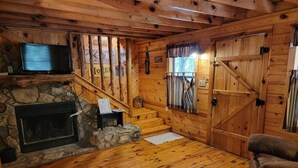 A wooden cabin interior with a stone fireplace, a TV mounted above it, a staircase leading to a loft, and a large wooden door.