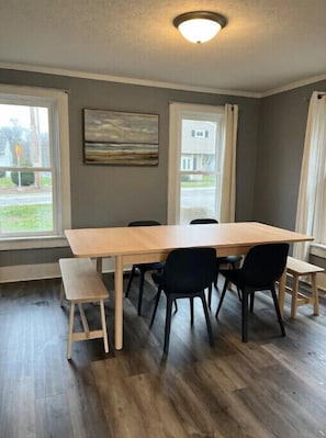 Un comedor con una mesa y sillas de madera, ventanas amplias y un cuadro en la pared.