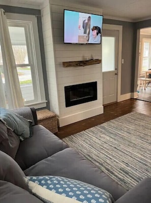 Una sala de estar con chimenea, televisor montado en la pared y un ventanal grande.