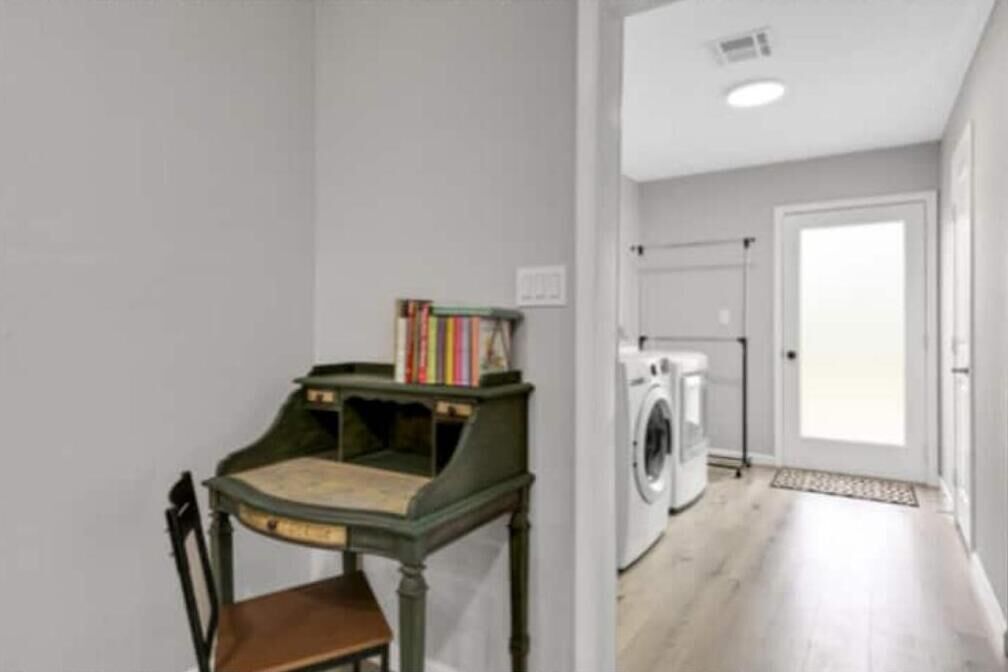 Una habitación con una pequeña mesa y una silla, una lavadora y una puerta que da a otra habitación.