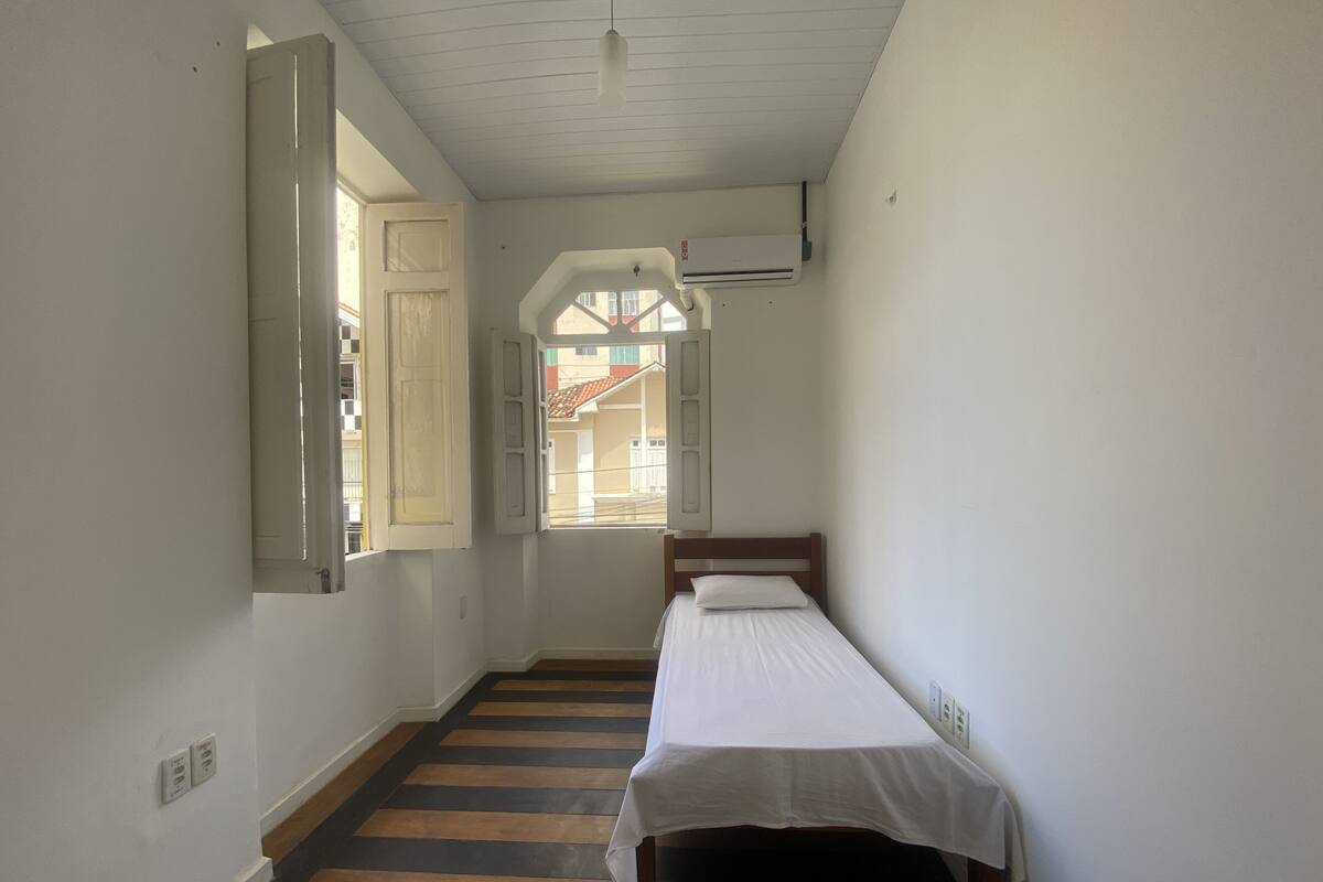 Ein schmaler Flur mit einem Bett am Ende, einem Fenster mit geöffneten Klappläden und einem Deckenventilator.