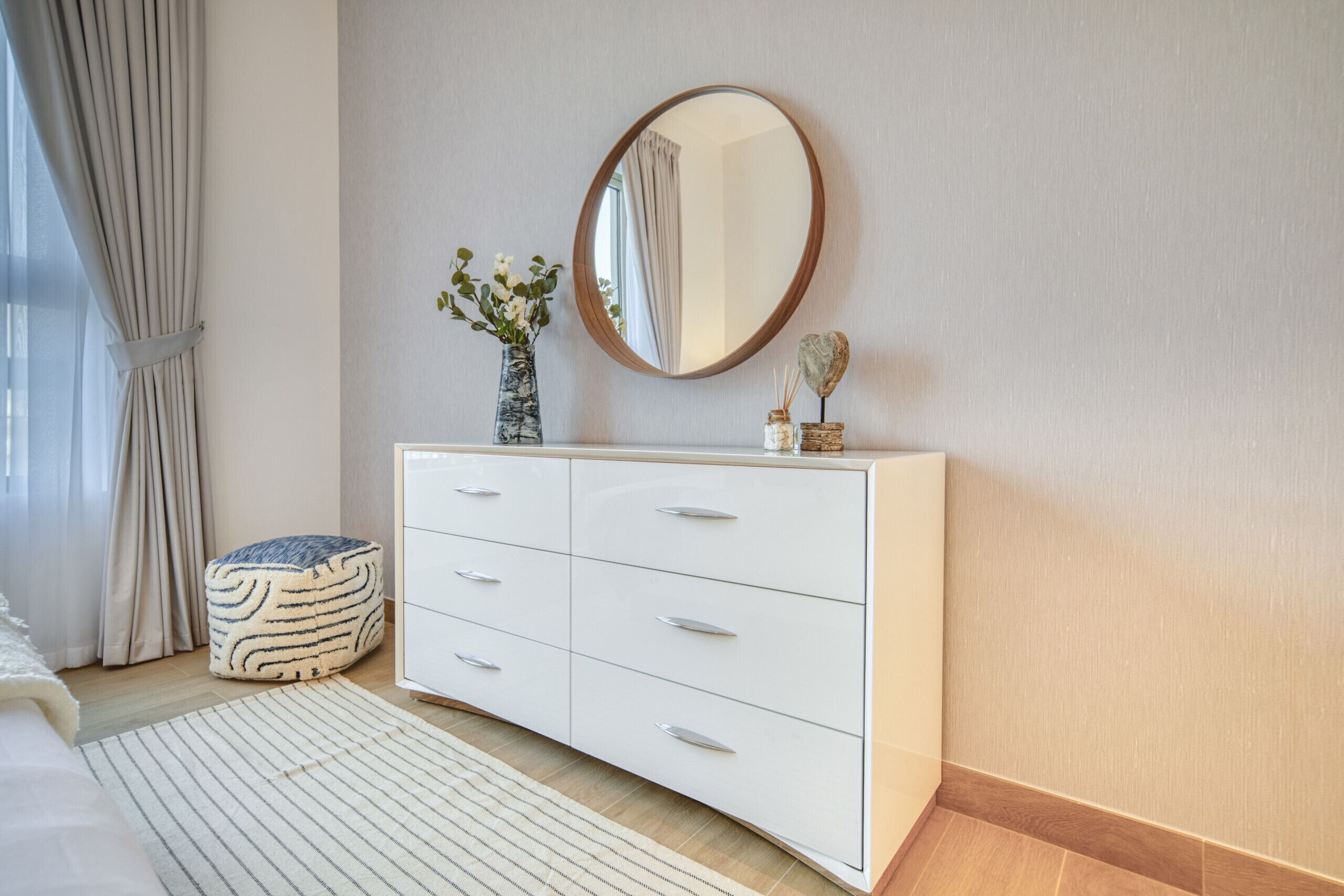 A modern bedroom with a white dresser, a round mirror, a vase with flowers, and a striped rug.