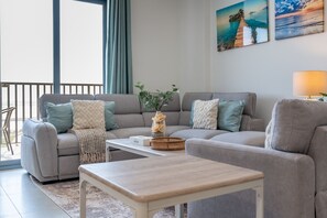 A living room with a grey sofa, a coffee table, and a lamp. There is a window with curtains and a view of the outside.