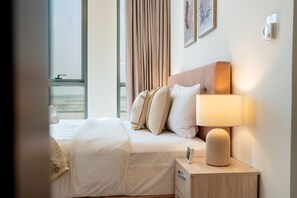 A hotel room with a bed, a bedside table with a lamp, a window with curtains, and a view of the beach.