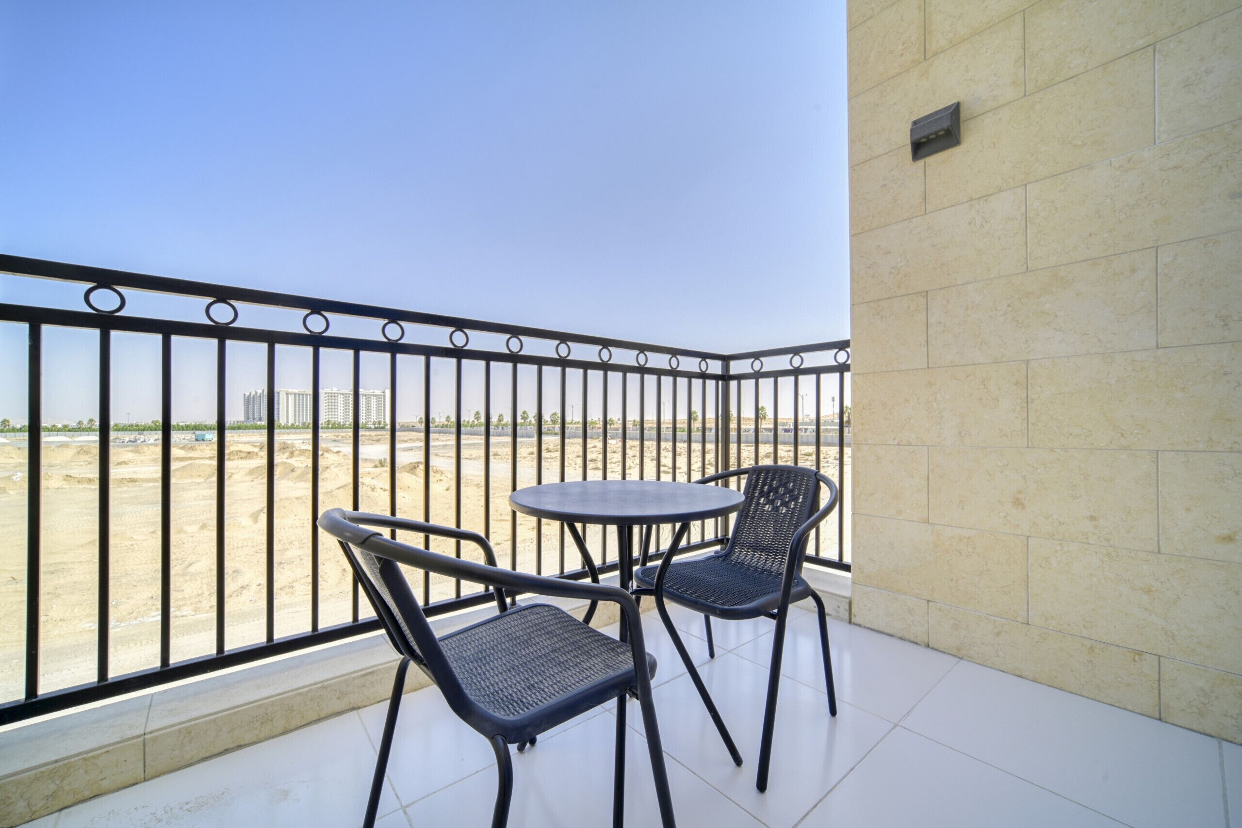 A balcony with two chairs and a table, overlooking a sandy area with buildings in the distance.