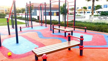 Ein Spielplatz im Freien mit einer hölzernen Bank, roten und blauen Spielgeräten und einem gelben Ball.