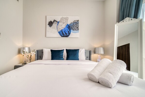 Un lit bien fait, avec du linge de lit blanc et des coussins bleus, un tableau au-dessus et un miroir au mur.