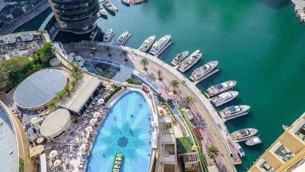 Vista aerea di un porto turistico con barche ormeggiate, una piscina con motivi geometrici e una passeggiata con lettini e ombrelloni.