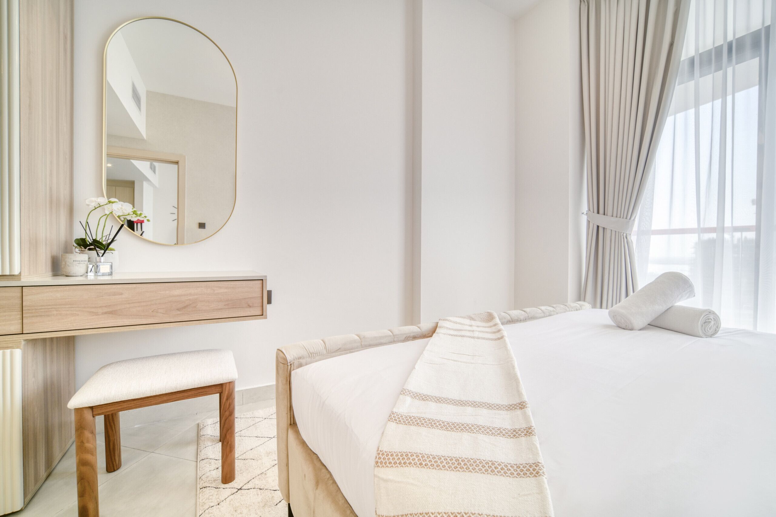 Une chambre d’hôtel moderne avec un grand lit, un banc en bois, une table de chevet ornée d’un vase de fleurs et un grand miroir au mur.