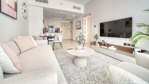 Un salon moderne avec un canapé, une table basse et un téléviseur. On aperçoit une cuisine au fond.