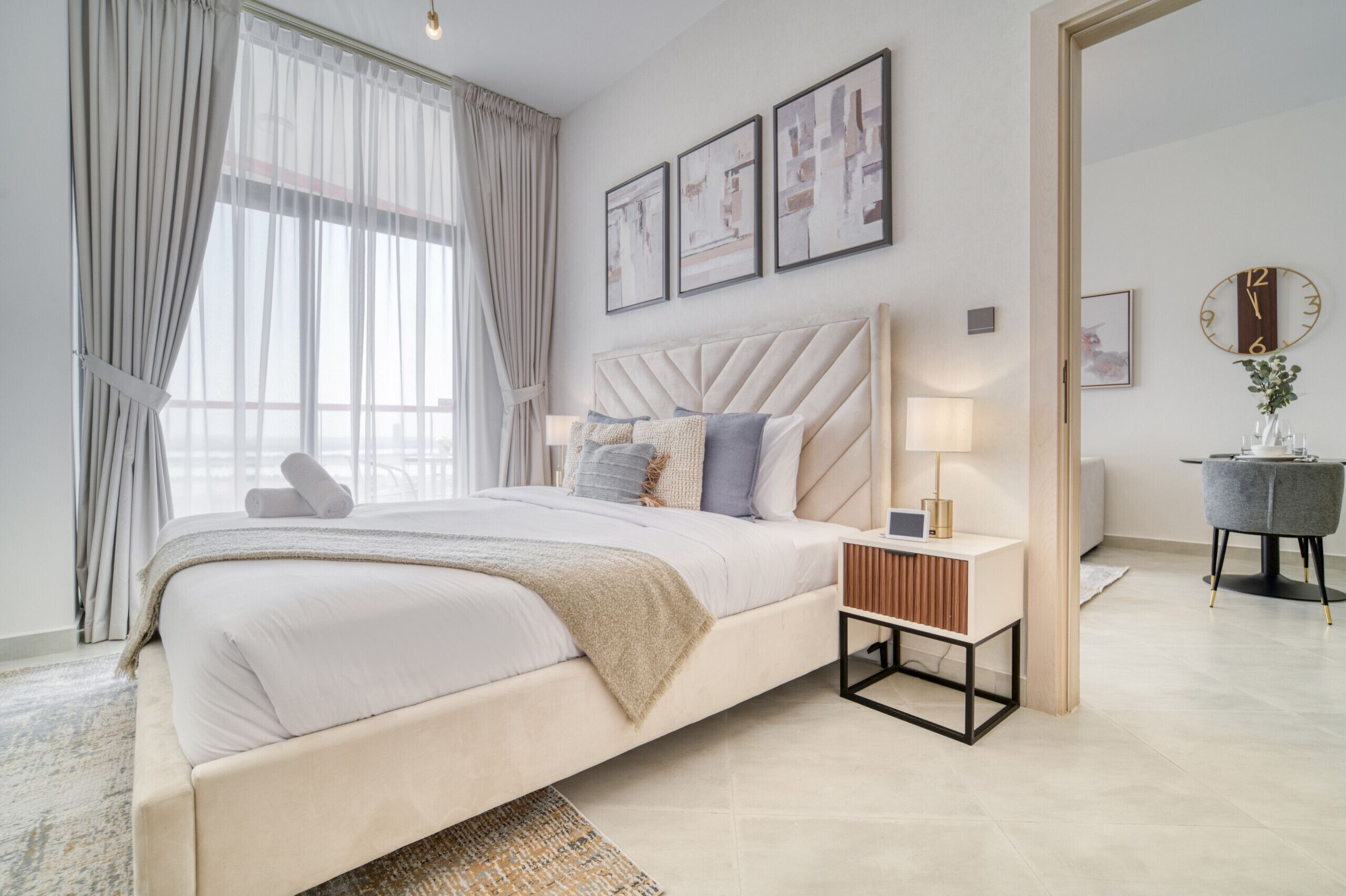 A modern bedroom with a large bed, a bedside table with a lamp, and a view of the outside through sheer curtains.