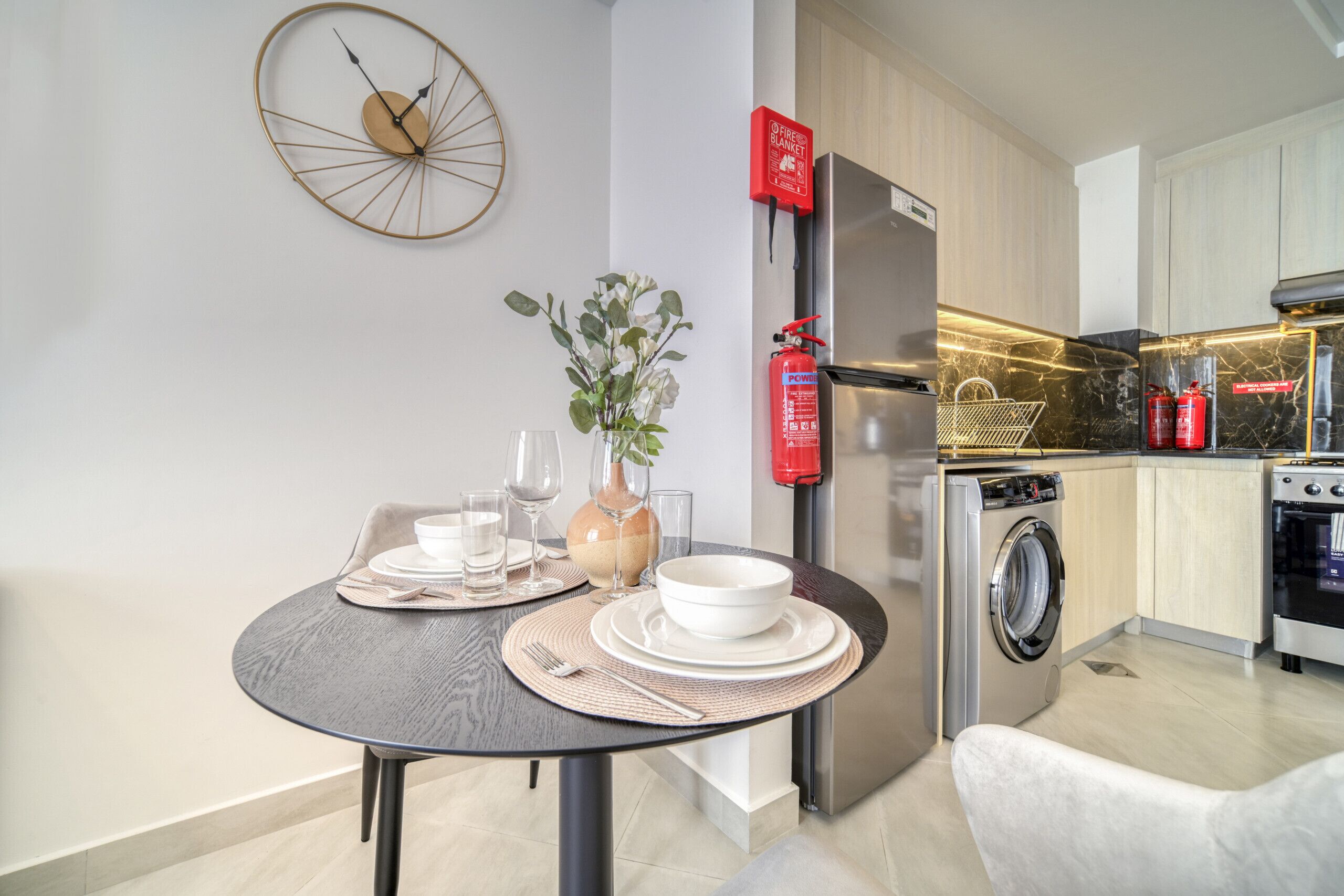 Una cocina moderna con lavadora incorporada, un extintor y un comedor con mesa puesta para dos.