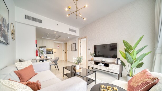A modern living room with a sofa, coffee table, and a TV. There is a kitchen in the background and a large plant. - Spectacular 1BR at Binghatti Avenue Al Jaddaf by Deluxe Holiday Homes (Dubai)