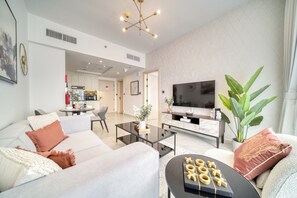 A modern living room with a sofa, coffee table, and a TV. There is a kitchen in the background and a large plant.
