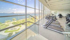 Ett gym med löpband och annan träningsutrustning, stora fönster med utsikt över en sjö eller ett hav och stadens skyline.