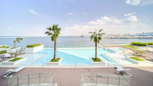 Ein Poolbereich auf dem Dach mit Palmen, Liegestühlen und Blick auf das Meer und die Skyline der Stadt.