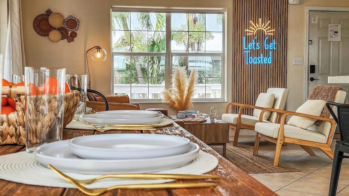 A dining area with a long wooden table set for a meal, a neon sign reading "Let's Get Toasted," and a view of palm trees through large windows.
