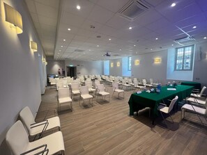 Egy konferenciaterem, sorokban elrendezett székekkel, egy hosszú asztallal és egy projektorvásznon.