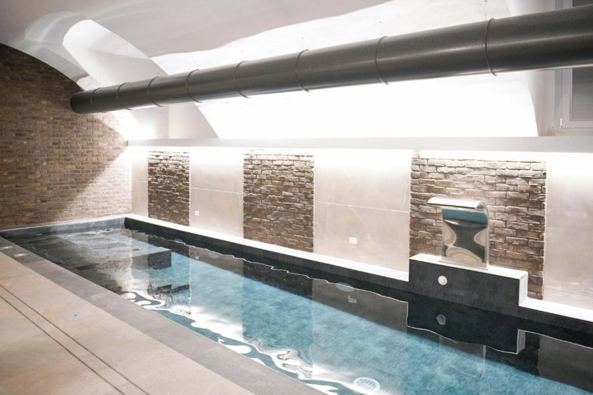 A modern indoor swimming pool with a clear water surface, a brick wall on one side, and a stone wall on the other.
