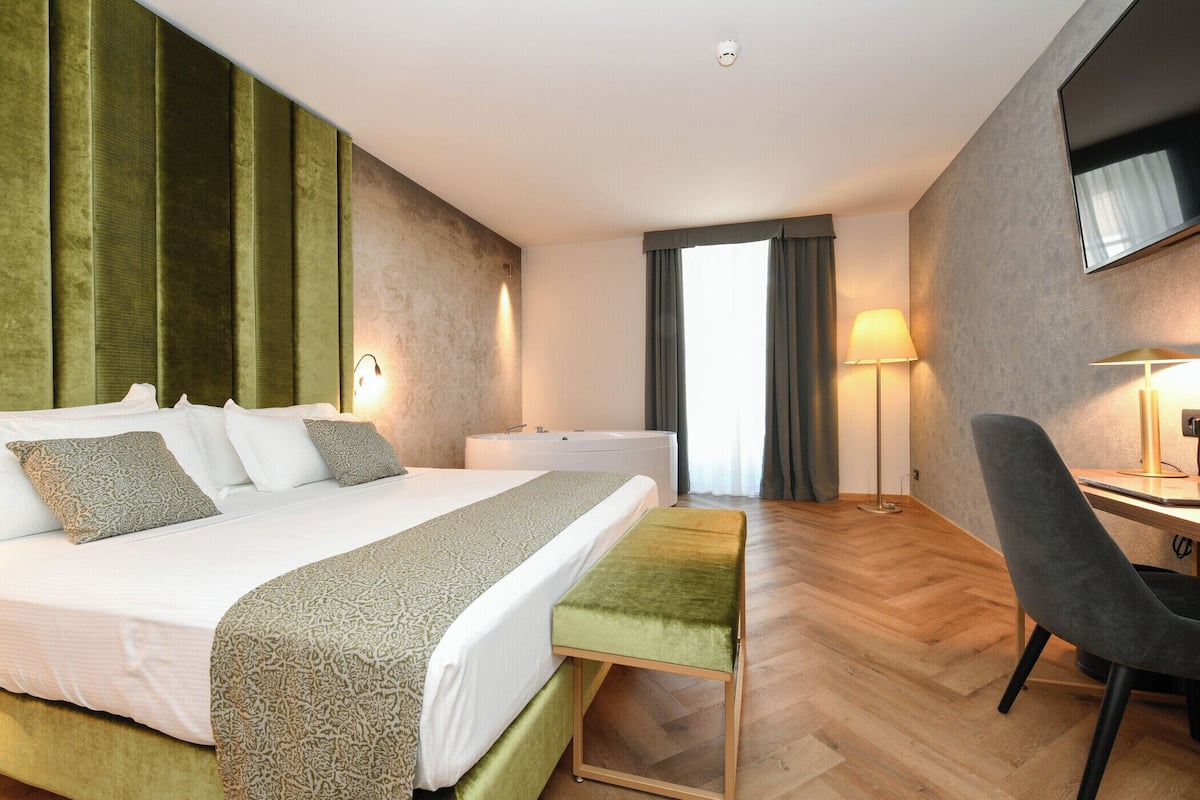 A hotel room with a bed, bedside table, desk, chair, and a large window.