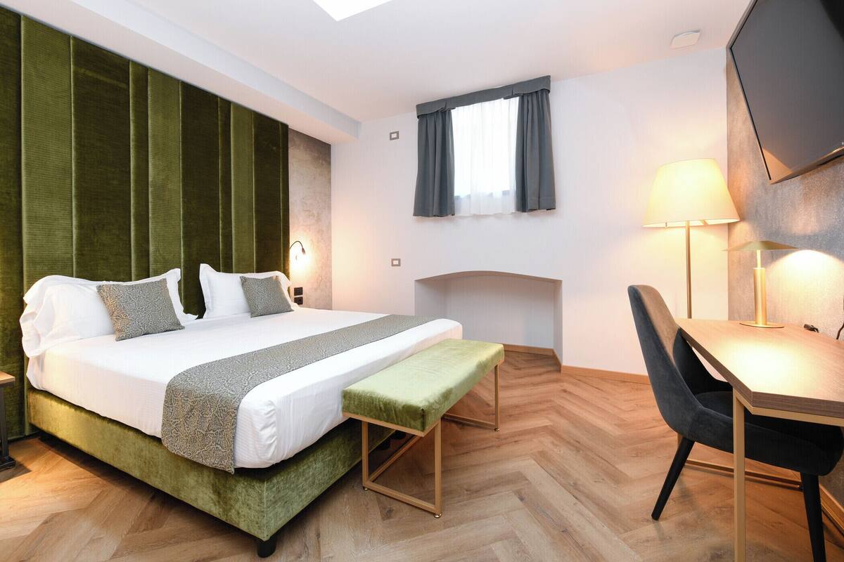 A modern hotel room with a large bed, a wooden desk, and a green bench.