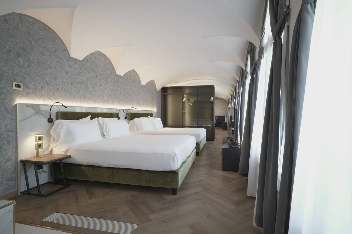A hotel room with two beds, a nightstand, a lamp, and a large window with curtains.