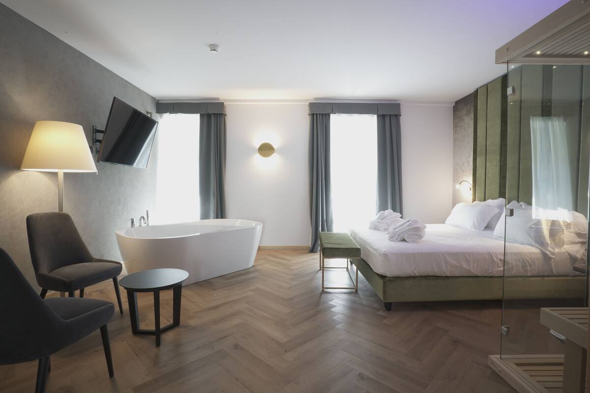 A modern hotel room with a large bed, a sitting area with a chair and a small table, a bathtub, and a flat-screen TV mounted on the wall.