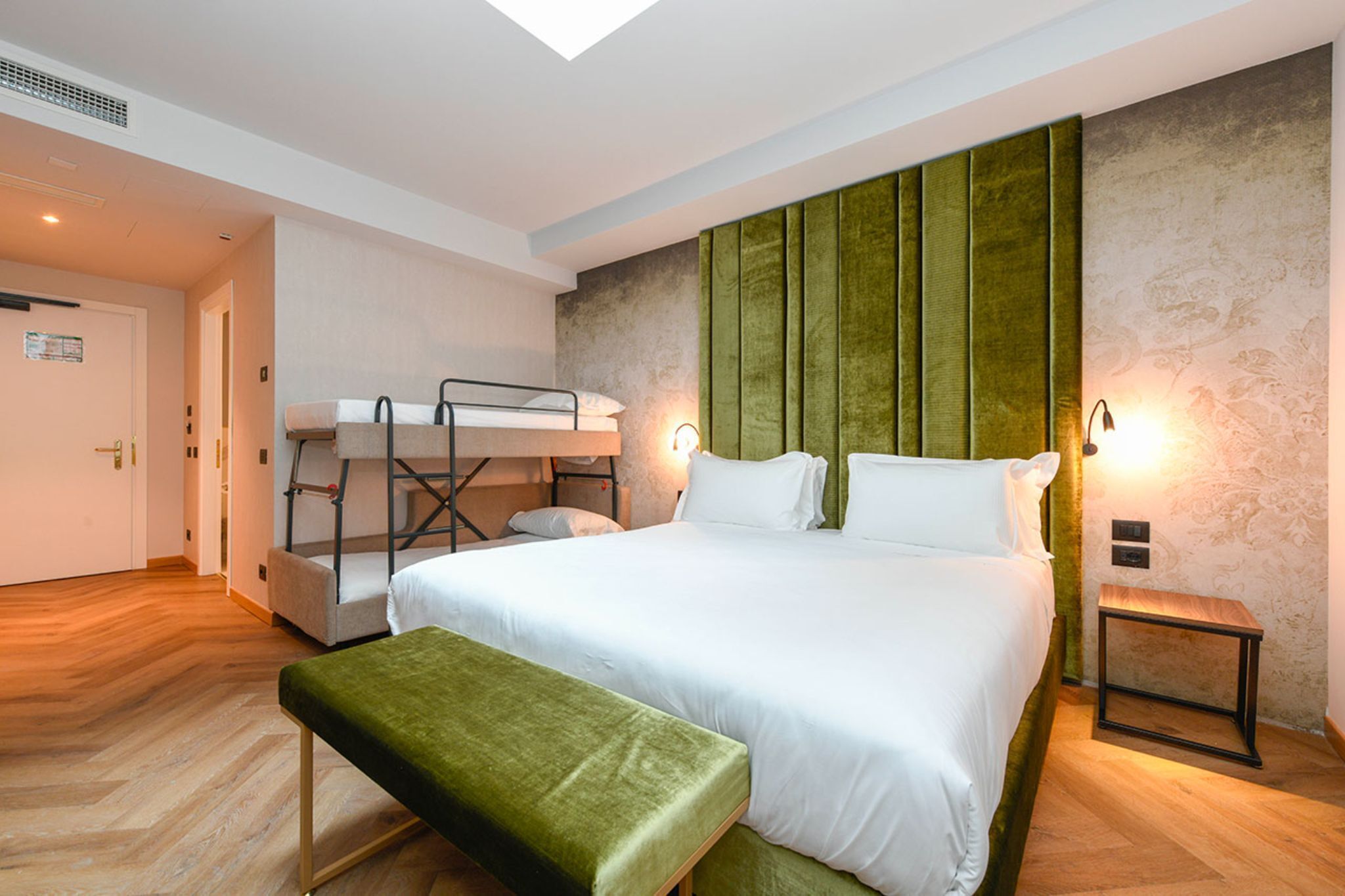 Una camera d'albergo con un letto a castello, un letto singolo, una panca verde e un tavolino.