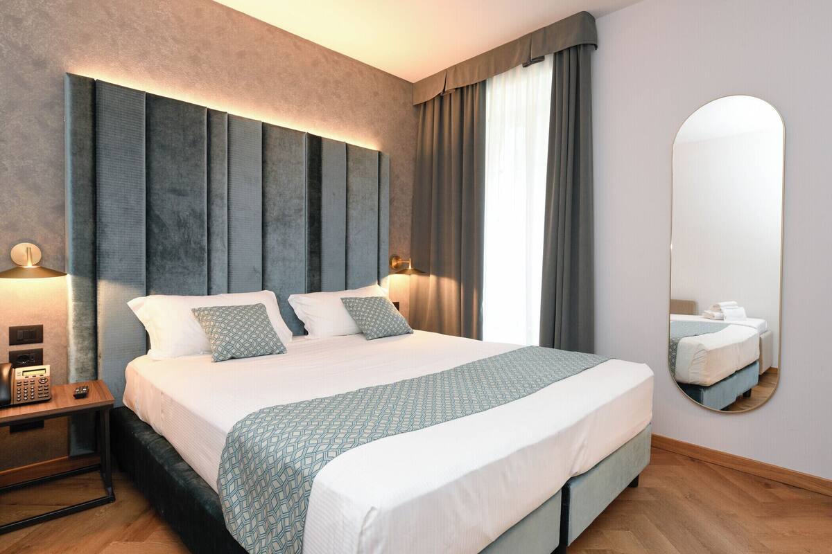 A modern hotel room with a large bed, a bedside table with a lamp, and a large mirror on the wall.
