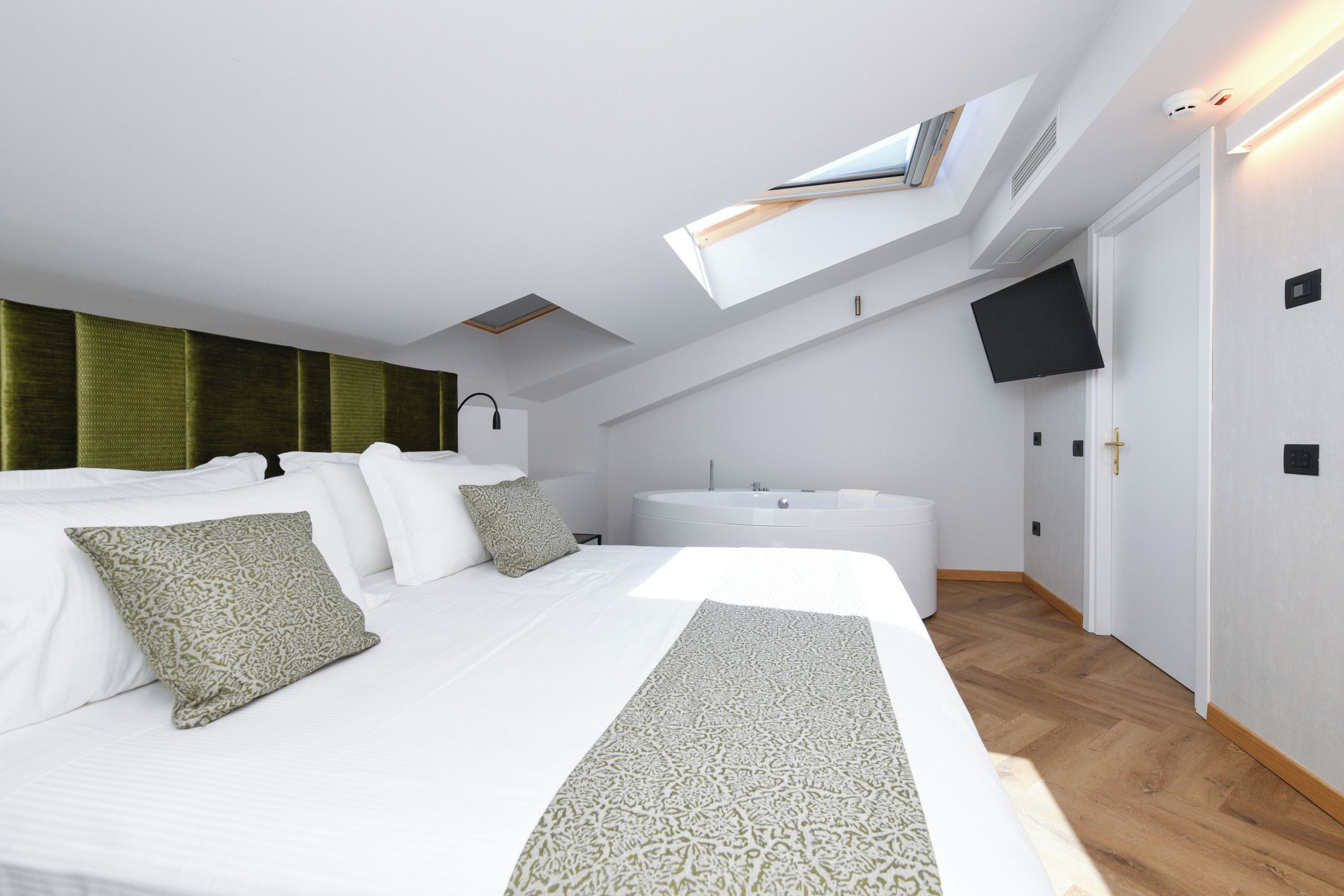 A modern bedroom with a large bed, a skylight, a bathtub, and a flat-screen TV.