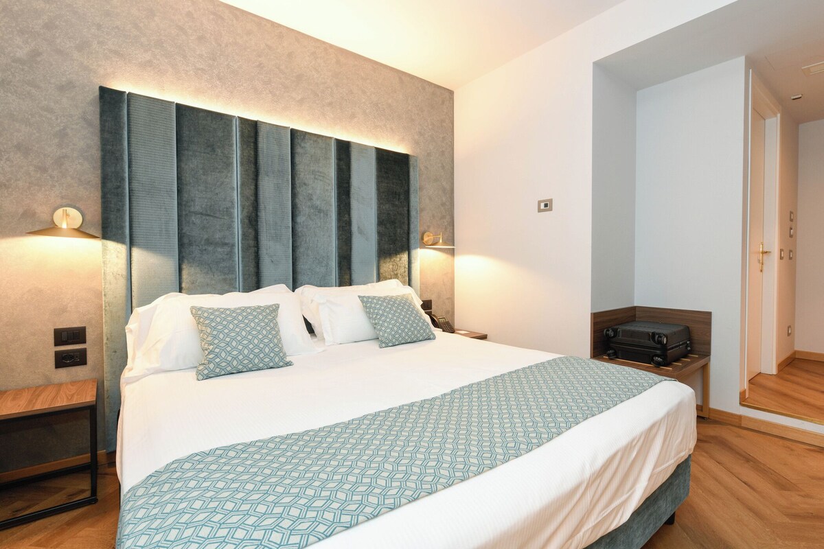 A neatly made bed with a geometric patterned bedspread, a headboard with vertical panels, bedside lamps, and a suitcase on a wooden nightstand.