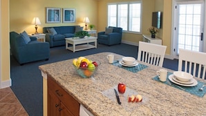 A living room with two blue sofas, a coffee table, and a dining area with a granite countertop, plates, and a bowl of fruit. - Three Bedroom, Luxury Condo, Hilton Head Island, SC 3348165 (Hilton Head Island)