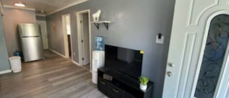 Un pasillo con ventilador de techo, refrigerador, televisor sobre un soporte negro y una puerta con panel decorativo.